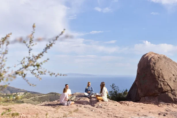 Four adults meditate on a rocky cliff overlooking the ocean under a clear, sunny sky.
