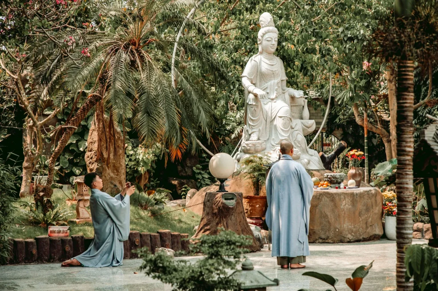 Buddhist monks meditating by a statue in a serene garden setting.