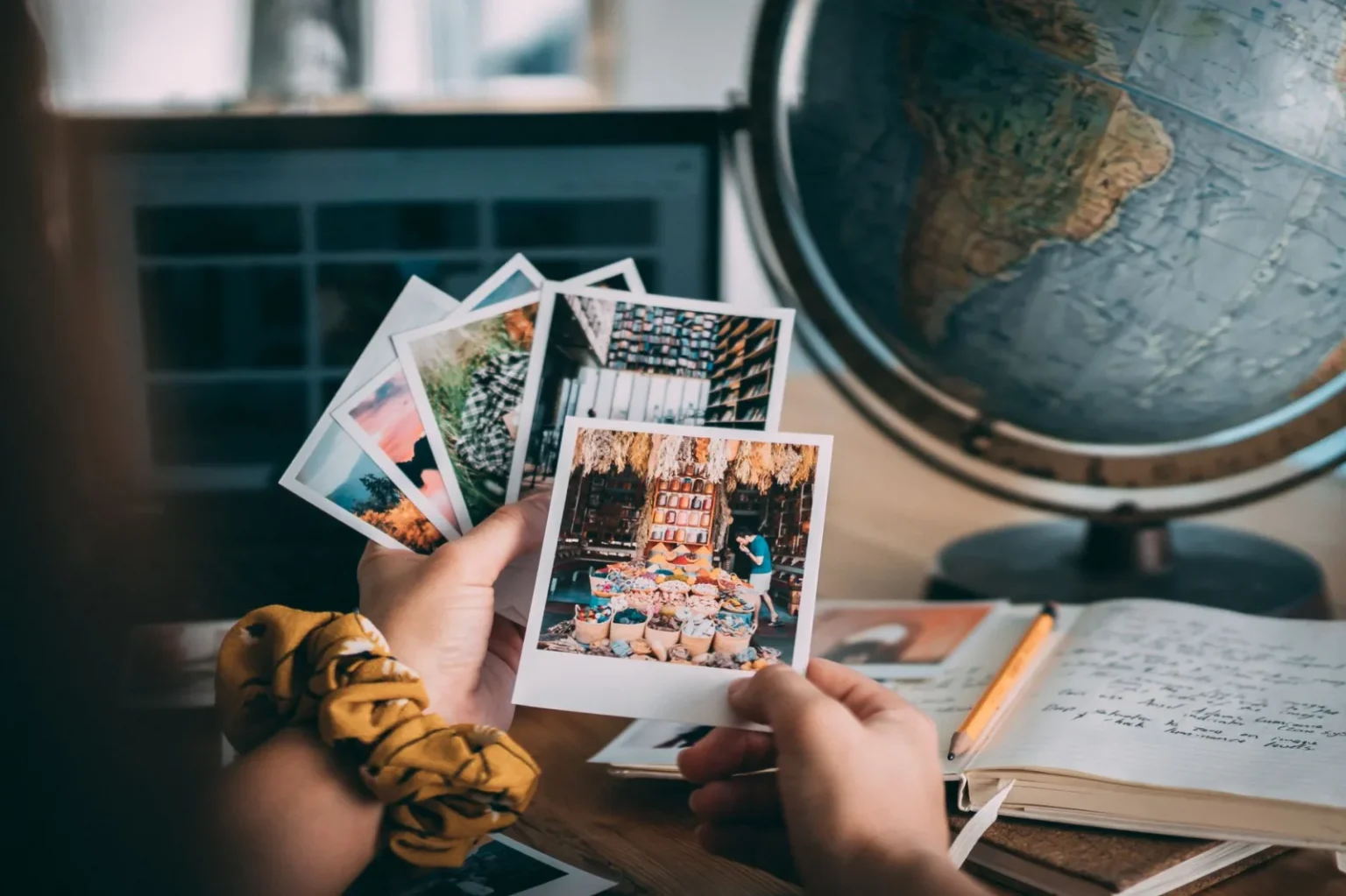Hands holding Polaroid photos with a globe and journal, evoking travel memories.