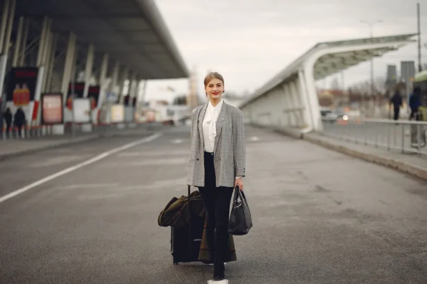Full body of young woman in stylish outfit carrying suitcase and bag while walking along pavement near modern airport building